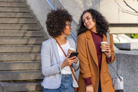 Diverse women chatting with coffee and smartphone on city steps, business casual lifestyle