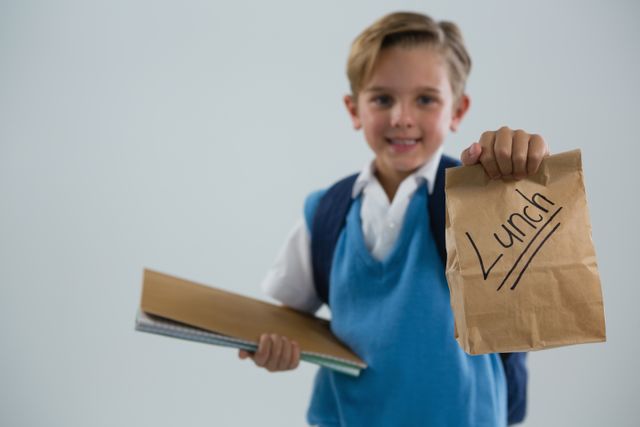 Smiling schoolboy holding book and lunch paper bag against white background