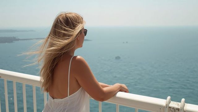 Blonde woman wearing sundress and sunglasses gazing over ocean from sunny terrace