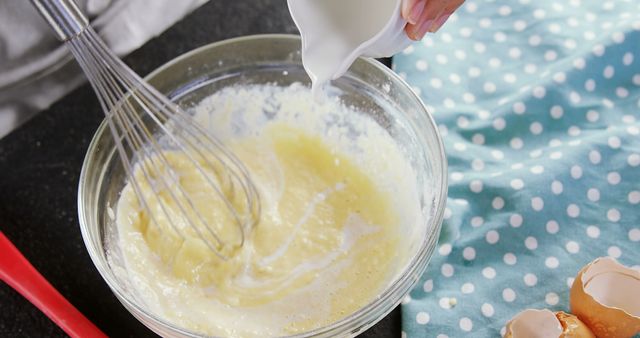 This stock image showcases the process of whisking batter in a glass bowl while adding milk. The homemade, hands-on approach in the kitchen scene is highlighted by the presence of eggshells and a polka dot napkin, making it perfect for illustrating baking recipes, cooking websites, culinary blogs, or DIY cookbook content.