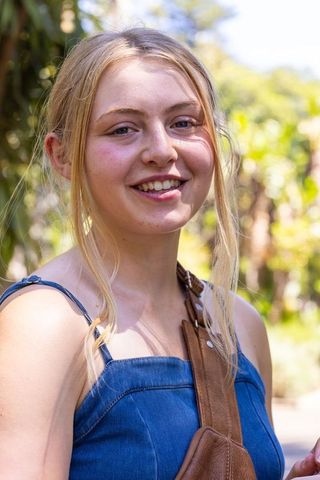 Smiling young woman in sunlit park wearing denim top and brown leather crossbody