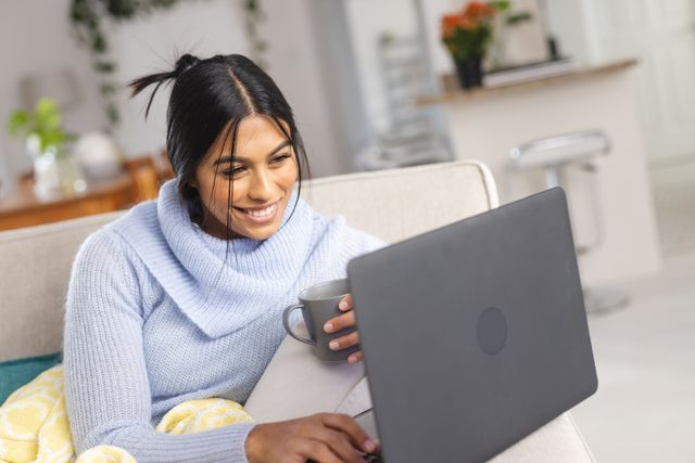 Smiling middle eastern young woman with coffee cup video calling through laptop at home. Unaltered, lifestyles, communication, wireless technology, refreshment, online.