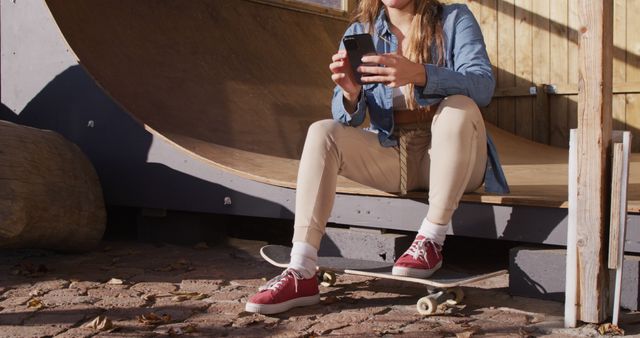 Image of happy caucasian female skateboarder resting and using smartphone. Skateboarding, sport, active lifestyle and hobby concept.