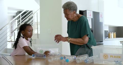 African grandmother teaching granddaughter recycling with plastic bottles in modern kitchen