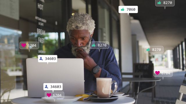 Man with gray hair and beard seated at outdoor café table with laptop and coffee, surrounded by digital icons representing social media engagement, like hearts, thumbs up, and user icons. Scene conveys theme of connectivity, modern communication, and social engagement. Perfect for content about remote work, digital marketing, lifestyle blogs, or tech-savvy senior demographics.