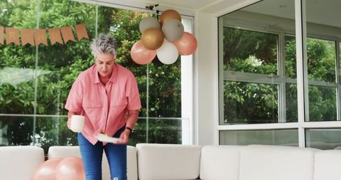 Senior woman tidying living room after celebration with balloons