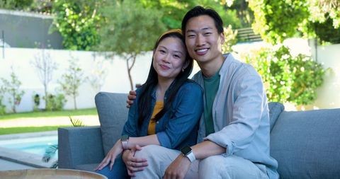Smiling couple relaxing on backyard sofa with smartwatches poolside and greenery