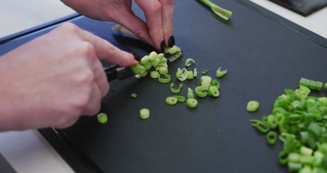 Hands expertly chopping green spring onions on a black cutting board, commonly used in cooking magazines, kitchen recipe books, food blogs, and culinary guide articles. A great visual for tutorials on food preparation, kitchen safety demonstrations, or recipe illustrations.