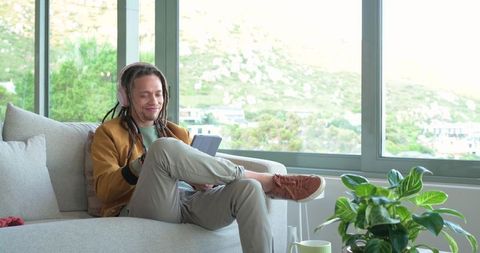 Mid-30s man relaxing on sofa wearing headphones and using tablet by bright window