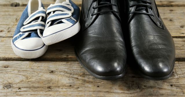 A pair of small blue and white sneakers is placed next to a pair of adult-sized black leather shoes on a wooden surface, with copy space. It symbolizes the contrast between childhood and adulthood, indicating a parent-child relationship or the concept of growth.
