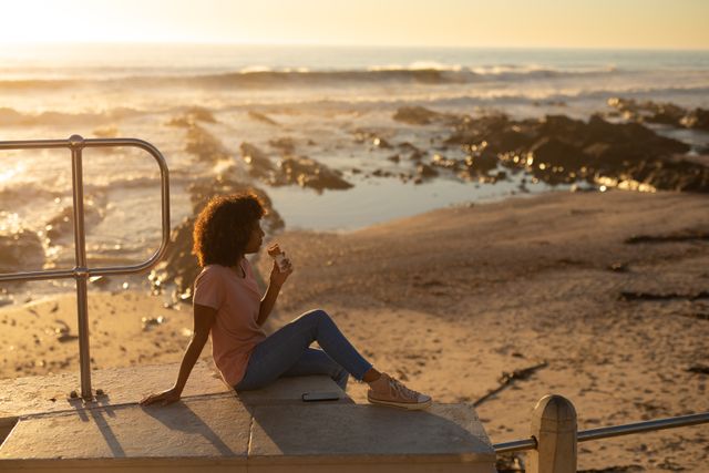 Side view of a mixed woman sitting by a fence on a promenade enjoying an ice cream on a sunny day by the sea, backlit by the setting sun