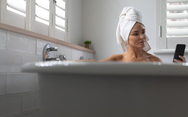 Woman sitting in a bathtub with a towel wrapped around her head, using a mobile phone. Ideal for themes related to relaxation, self-care, modern lifestyle, home comfort, and the integration of technology into everyday routines.