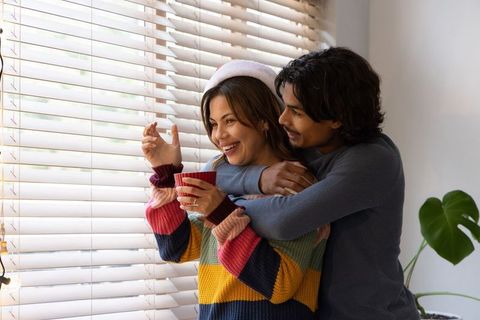 Cozy holiday couple embracing at window holding mug and smiling through blinds