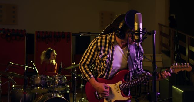 Front view of a biracial man with dreadlocks in a ponytail playing electric guitar and singing, and a blonde Caucasian woman playing drums together in a live recording room at a recording studio. Musicians working on producing a song