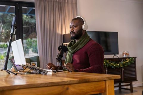 African american man recording podcast at home wearing headphones and scarf with laptop