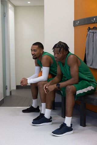Two teammates sitting on bench wearing green basketball jerseys holding basketball