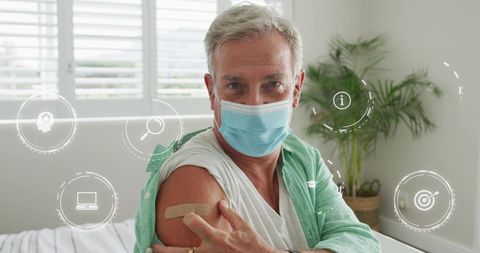 Senior man wearing mask showing vaccine bandage at home highlighting post-vaccine wellness