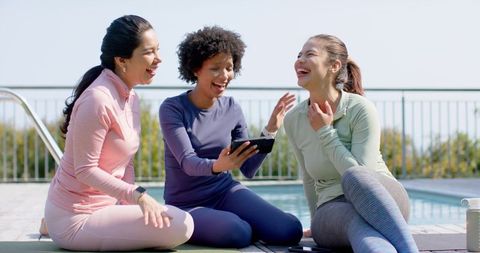 Diverse women laughing and sharing tablet on pool deck in pastel activewear for wellness