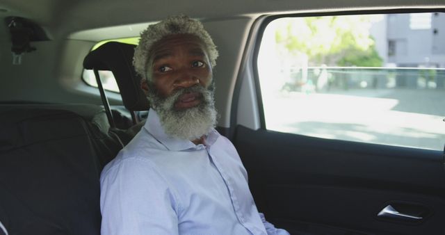 Elderly African American man with gray hair and beard is sitting in the backseat of a car and looking out the window with a thoughtful expression. He is wearing a light blue shirt. This can be used in contexts related to transportation, introspection, ride services, travel, road trips, senior lifestyle, or older adults and mobility.