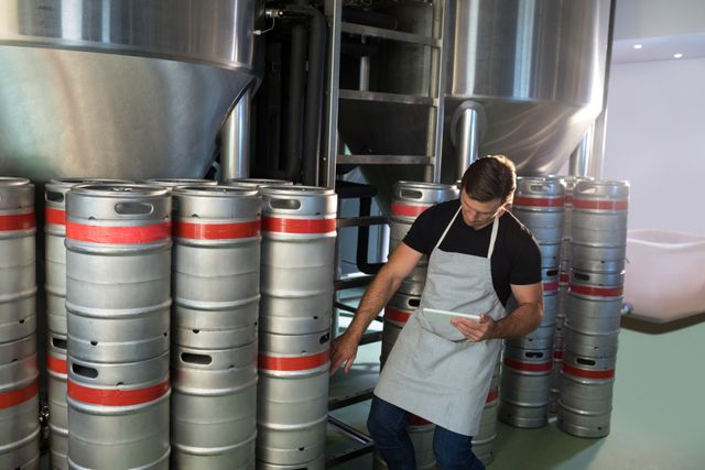 Worker in brewery warehouse counting kegs using a tablet. Ideal for illustrating industrial processes, inventory management, and modern technology in traditional industries. Suitable for articles on brewery operations, logistics, and production efficiency.