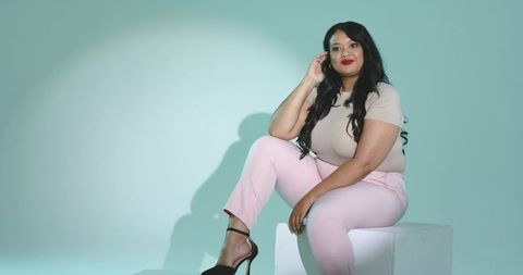 Confident mid-adult woman posing on cube wearing pink trousers and black heeled sandals