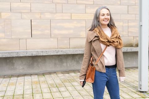 Mature woman walking and smiling in urban courtyard wearing tan coat and leather crossbody