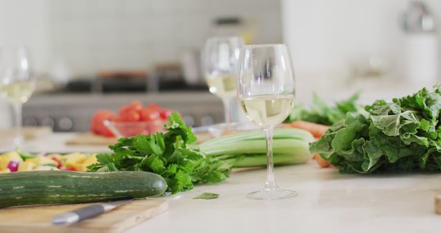 This image showcases fresh vegetables like cucumber, parsley, celery, and leafy greens arranged on kitchen countertops with a wine glass, suggesting a healthy lifestyle. In the background, tomatoes and other foods are out of focus. Ideal for use in healthy eating articles, cooking blogs, kitchen equipment advertisements, or wine pairing promotions.