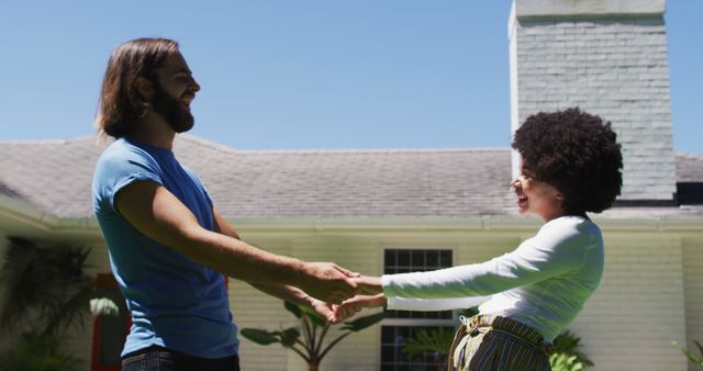 A cheerful couple enjoying a bright sunny day outdoors, holding hands and smiling at each other in front of a house. Perfect for themes related to relationships, happiness, outdoor activities, home lifestyle, diversity, and joy. Ideal for use in blog posts, social media content, advertisements, and articles focusing on love, interpersonal bonds, and lifestyle content.