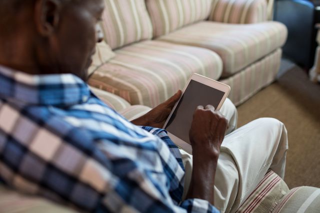Senior man sitting on a sofa at home using a tablet computer. Ideal for illustrating concepts related to elderly people embracing technology, digital communication, modern lifestyle, and leisure activities at home.