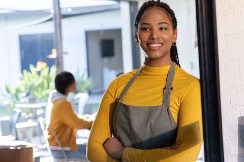 African american barista smiling in sunlit cafe wearing gray apron and mustard top