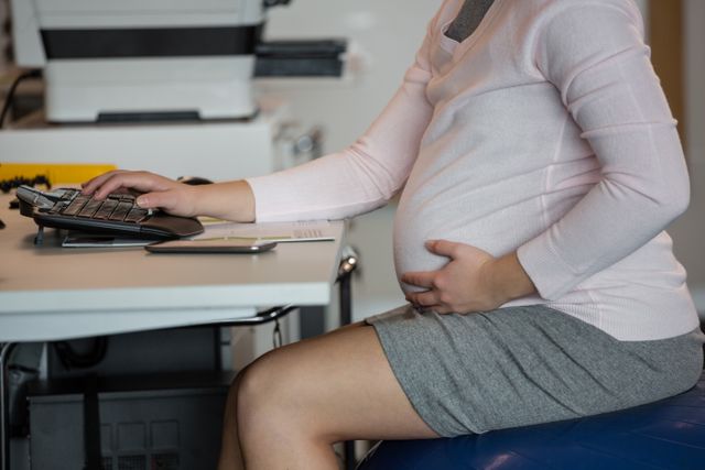 Pregnant businesswoman working at her desk, typing on a computer keyboard. Ideal for use in articles or advertisements related to maternity in the workplace, work-life balance, professional women, and corporate environments. Can also be used in blogs or websites focusing on pregnancy, career advice, and women's health.