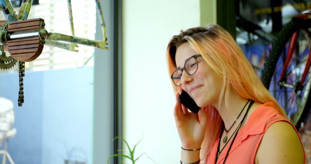 Smiling young woman with pink hair and glasses talking on phone in bicycle shop. Appropriate for themes related to communication, technology, modern lifestyle, customer service, retail, and casual interactions. Useful for websites, blogs, marketing materials, and advertisements.