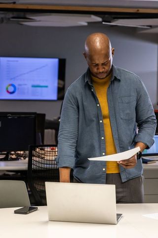 African american man reviewing reports at office desk with laptop and charts for analytics