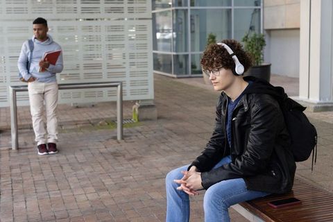 Diverse college students listening to music and waiting on campus bench with backpack