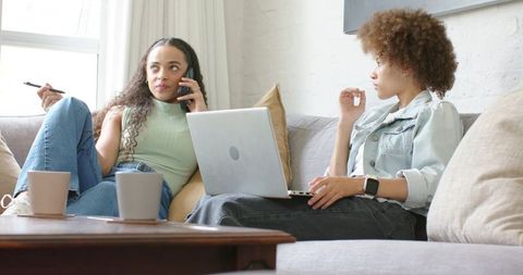 African american women working remotely and chatting on sofa with laptop and phone