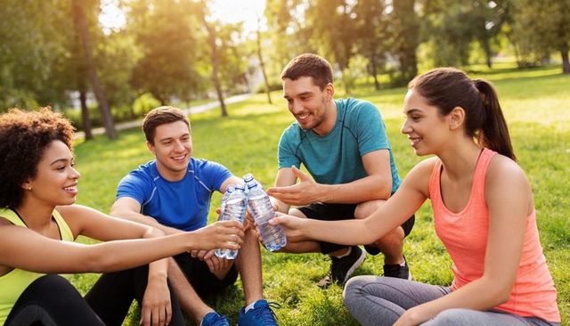 Friends toasting water bottles after run in park during golden hour, fitness recovery
