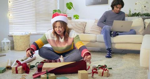 Cozy holiday scene asian couple wrapping christmas gifts at home with man working on laptop