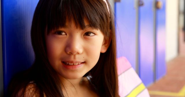 Smiling young girl with long hair and headband sitting close to brightly colored school lockers. Scene captures the essence of childhood and school life, full of brightness and carefree attitude. Ideal for educational websites, school advertisements, children's programs, and youth-centric marketing materials.