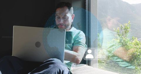 Focused man working on laptop by window with digital globe overlay and coffee mug