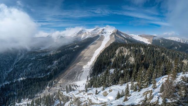 Snowy central ridge rising over conifer forest with avalanche chute and dramatic sky