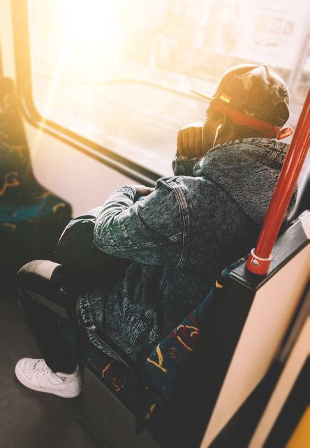 Man seated on public transport in morning light wearing denim jacket and headphones, gazing out window. Useful for themes involving commuting, city life, solo travel, or reflective mood. Can be used in advertisements for travel, urban lifestyle promotions, or public transportation announcements.