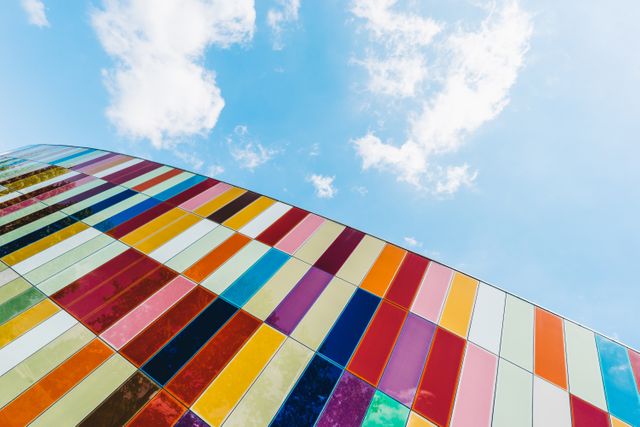 Colorful glass façade of a modern building with vibrant panels against a clear blue sky with white clouds. Perfect for use in advertising for urban development, architectural designs, or promoting modern city living. Can also be used for backgrounds, websites, or art-related content.
