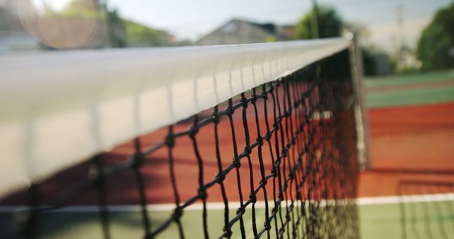 Photo captures a detailed, close-up look at a tennis net on a sunlit outdoor court. Ideal for use in sports-related content, blogs about tennis, recreational activities, or websites promoting health and fitness. The image could also be used to highlight the textures and materials involved in sports equipment.