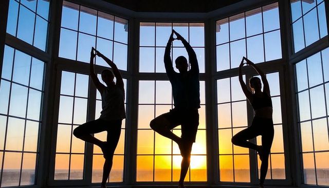 Balancing silhouetted adults practicing yoga at sunset by large bay window