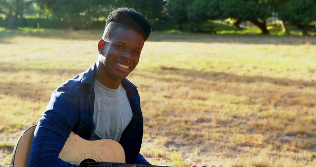 Young man playing guitar and smiling in a grassy field. This image is perfect for themes related to music, relaxation, nature, and leisure activities. Ideal for use in lifestyle blogs, music websites, and promotional material for outdoor events or youth programs.