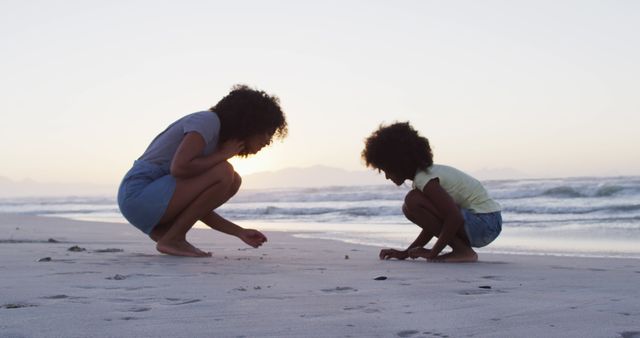 Mother and daughter spending quality time by beach at sunset, kneeling and inspecting sand. Perfect for use in promotions for family vacations, beach resorts, and moments of bonding between parents and children.