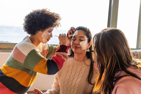 Three friends applying face masks on sofa by sunlit window wearing cozy knitwear