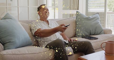 Middle-aged man relaxing on sofa holding remote watching tv in bright modern living room
