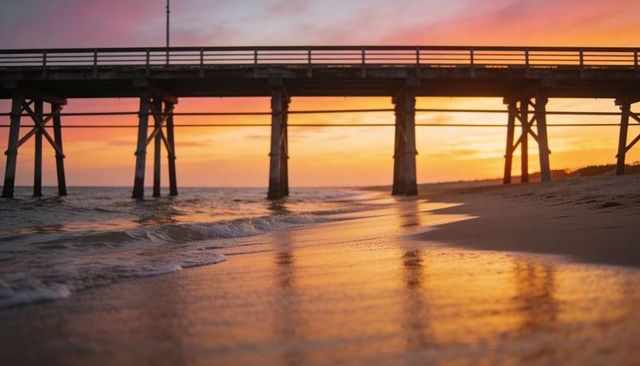 Golden-hour pier reflecting on wet sand with silhouetted pilings and pastel sunset sky