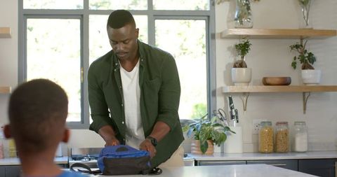 African american father packing boy's blue backpack on kitchen counter while son watches